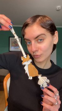 Portachiavi a forma di volpe color sabbia e bianca con un nappa bianca; realizzato in materiale simile alla pelle, presenta cuciture dettagliate e un'espressione giocosa.