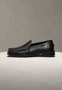 Mocassin en cuir noir avec bout moc cousu, talon bas et petite étiquette logo en tissu sur le côté, présenté sur une surface et un fond neutres.