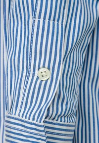 Modrobíle pruhovaná košile s knoflíkovým zapínáním, vyrobená z bavlněné látky; má dlouhé rukávy a manžetu s viditelným prošitím.