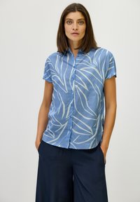 Camicetta azzurra chiaro a maniche corte con un motivo a foglie bianche, davanti bottonato e un fit rilassato, abbinata a pantaloni scuri.