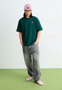 Tricou din bumbac verde, supradimensionat, cu un mic logo roșu, blugi gri cu croi larg, o căciulă roz și adidași roz, așezate pe un podea verde.