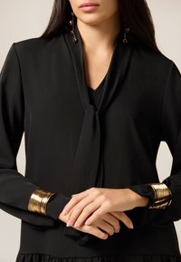 Zwarte blouse met een striksluiting bij de hals, lange mouwen en gouden metalen manchetten. Soepele textuur met een vloeiend ontwerp. Subtiele V-hals.
