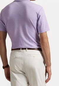 Uomo che indossa un polo a righe viola chiaro con maniche corte infilato in pantaloni avorio, con una cintura in pelle marrone e un orologio al polso, visto da dietro.