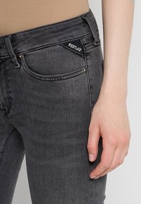Gros plan sur une personne portant un jean Replay gris foncé, montrant la poche avant et une partie de la main reposant à côté de la cuisse.