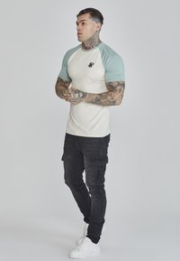 Men's short-sleeve, cream and mint green raglan shirt with a small black logo, paired with black cargo pants and white sneakers.