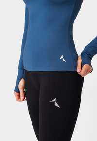 Zils garām piedurknēm tops no gludas auduma, savienots ar melniem ribotiem legingiem. Abi satur atstarotājus ar logotipiem. Tuvplānā redzams pieguļošs dizains un auduma tekstūra.