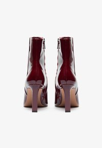 Botines de piel charol burdeos con un diseño elegante, punta afilada, acabado brillante y tacones de aguja de 10 cm; cierre con cremallera lateral.