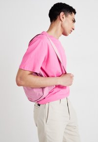 Rožinė audinio peties rankinė su lygiu paviršiumi, nedidelio dydžio, su reguliuojama dirželiu, dėvima kartu su derančiu rožiniu marškinėliu ir šviesių spalvų kelnėmis.