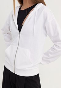 Sweat à capuche blanc à fermeture éclair, en tissu doux, doté d'une poche avant, d'une capuche avec cordon de serrage, et d'une taille côtelée. Porté par-dessus un haut noir.