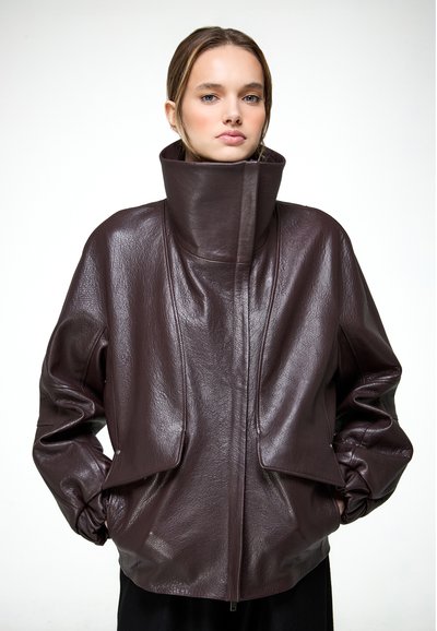 Žena oblečená v nadrozmernej tmavohnedej koženej bunde s vysokým, štruktúrovaným golierom, ruky vreckách, na bielom pozadí.