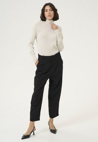 Helles beige Rollkragenshirt, schwarze hoch taillierte Hose und schwarze spitz zulaufende Absätze. Glatte Textur, maßgeschneiderte Passform und elegantes Design.