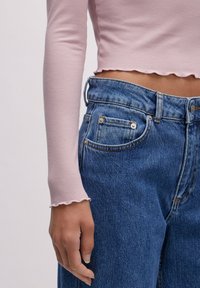 Gerippter, hellrosa Langarmtop mit gewelltem Saum, kombiniert mit hoch taillierten blauen Jeans aus Denim, die über zwei Vordertaschen und sichtbare Nähte verfügen.