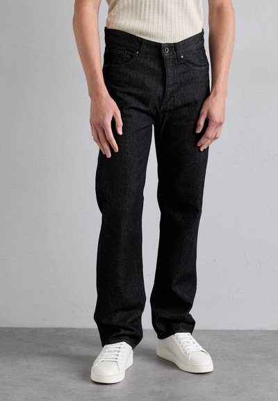 Jean en denim noir avec une coupe droite, comportant cinq poches, une fermeture à glissière et bouton, porté avec des baskets blanches.