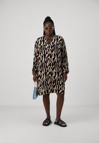 Robe à manches longues, longueur genou, avec un motif abstrait beige et noir. Tissu lisse, encolure ronde ornée de dentelle, associée à des sandales noires et un sac bleu.