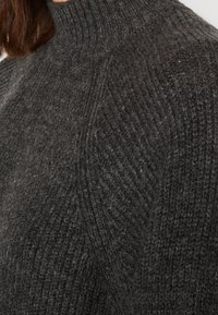 Primo piano di un maglione a collo alto di un grigio scuro, con texture a coste e motivi a treccia su spalle e collo.