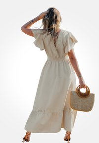 Robe maxi beige avec manches volantées, détails en dentelle à la taille et un lien à nouer dans le dos. Tient un sac à main rond en osier tressé.