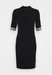 Robe noire longueur genou avec manches mi-longues, col montant, panneaux latéraux blancs et poignets de manches avec un texte noir audacieux.