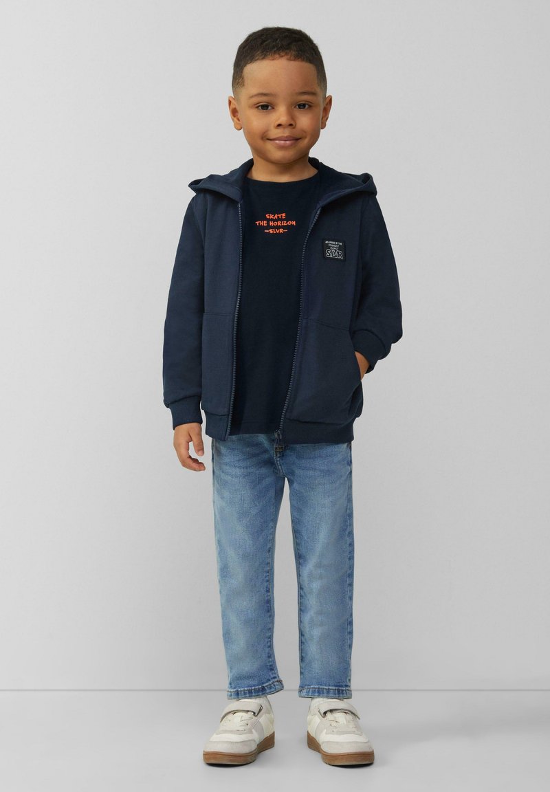 Marineblauwe zip-up hoodie met voorzakken over een donker shirt met oranje tekst, gecombineerd met lichtblauwe jeans en witte sneakers.