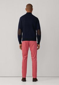 Marineblauer Strickpullover mit braunen Ellenbogenflicken, kombiniert mit rosa schmal geschnittenen Hosen und braunen Schuhen. Rückansicht vor einem schlichten Hintergrund.
