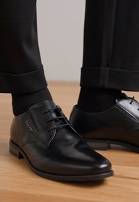 Schwarze Lederdressschuhe mit einem schlanken Design, versehen mit Schnürsenkeln, runder Schuhspitze und niedrigem Absatz. Getragen mit schwarzen Socken und maßgeschneiderten Hosen.