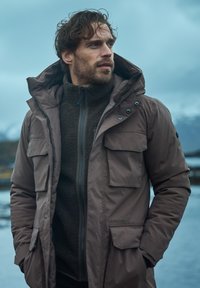 Brun vadderad parka med en pälsstruktur på insidan, hög krage och flera framfickor; har en blixtlåsförsedd front och justerbar huva.