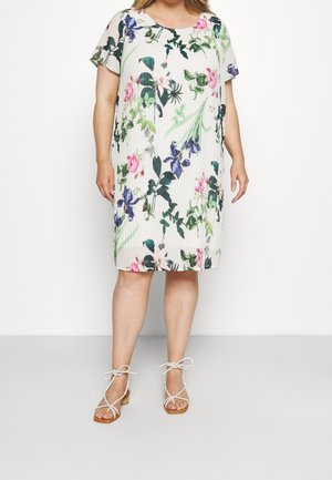 Femme portant une robe blanche mi-longue avec imprimé floral vert et violet et manches courtes, associée à des sandales blanches à brides.