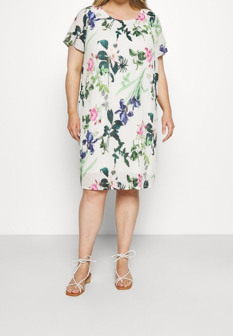Femme portant une robe blanche mi-longue avec imprimé floral vert et violet et manches courtes, associée à des sandales blanches à brides.