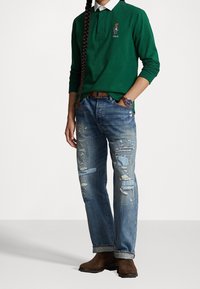 Person som bär en grön långärmad polo med björnlogotyp, slitna jeans med lappar, brun bälte, bruna mockaskor och en klocka på vänster handled.