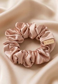 Chouchou en satin rose doux avec une petite étiquette beige indiquant "le siqúe" posé sur un fond de tissu lisse assorti.
