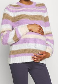 Kolorowy sweter w paski w odcieniach liliowym, beżowym i białym. Gruby splot, okrągły dekolt i ściągacze na rękawach. Noszony na szarych legginsach.
