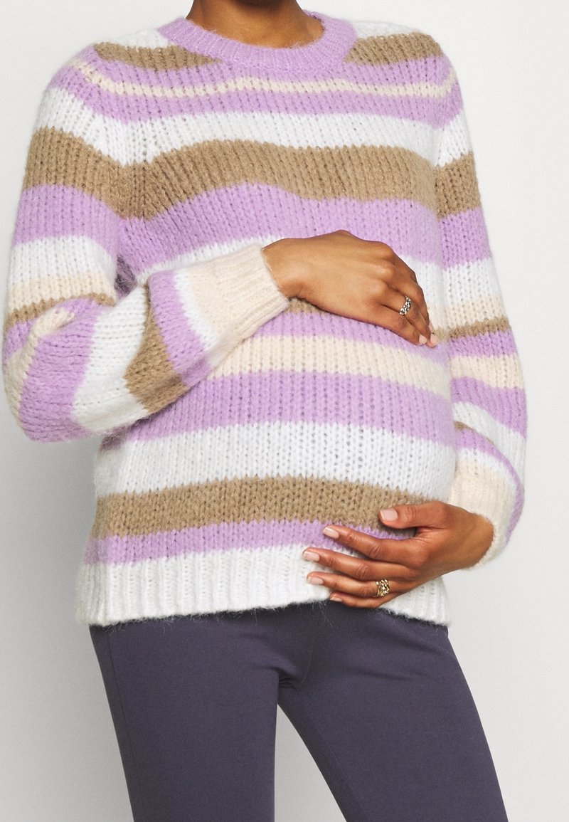 Kolorowy sweter w paski w odcieniach liliowym, beżowym i białym. Gruby splot, okrągły dekolt i ściągacze na rękawach. Noszony na szarych legginsach.