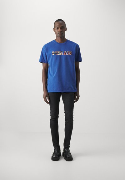 Camiseta de algodón azul con un gráfico rectangular multicolor en el pecho, combinada con jeans negros ajustados y zapatos negros.
