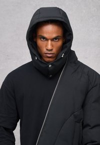 Giubbotto imbottito nero con colletto alto e cappuccio oversize, con zip diagonale e logo "Utopia" embossato sulla spalla.