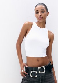 Biely, bezrukávový crop top s vysokým výstrihom, kombinovaný s čiernym širokým opaskom, ktorý má dve strieborné štvorcové spony. Čierne džínsy dotvárajú celkový vzhľad.