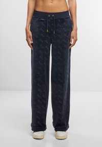 Navy velvet joggers with an all-over logo pattern, elastic waistband, adjustable drawstring, and straight leg fit. Paired with white sneakers.