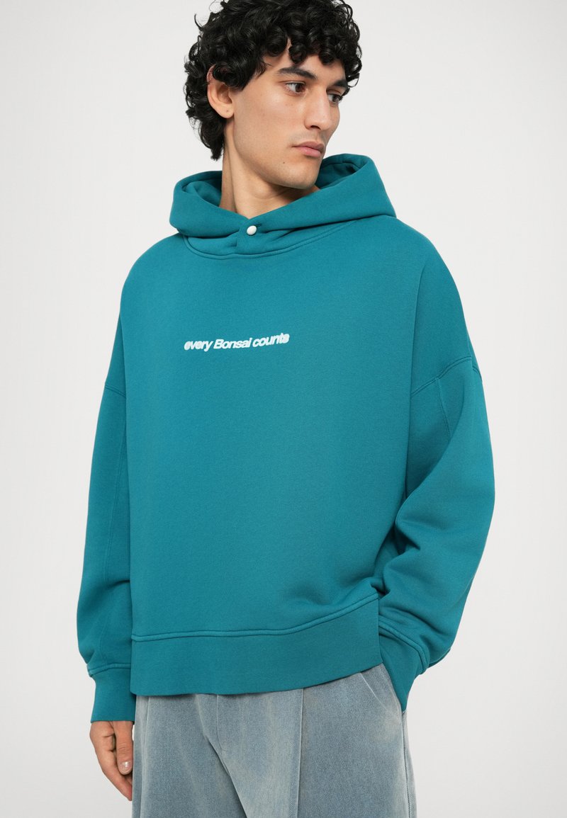 Türkise Kapuzen-Sweatshirt aus weichem Material, mit lockerem Schnitt und weißem Text "Jeder Bonsai zählt" auf der Vorderseite. Rippbündchen und Saum.