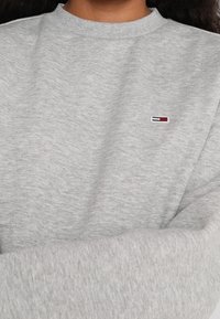Persona vestida con un sudadera gris claro que tiene un pequeño logo rojo, blanco y azul de Tommy Hilfiger en el pecho.