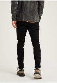 Svarta tajta byxor i kombination med en rutig långärmad skjorta. Byxorna har textur och bakfickor, och skorna är beiga med svarta accenter.