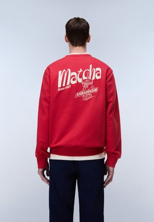 Mand iført rød sweatshirt med hvid tekst "Matcha Ambassadør Siden 1987" og tegneseriekrusmotiv, stående med ryggen mod, foran en ensfarvet baggrund.