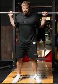 Homme en tenue de sport noire effectuant un squat avec barre dans une salle de sport avec un miroir et du matériel de musculation en arrière-plan.