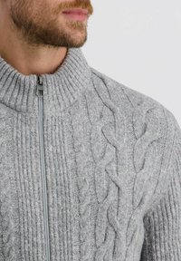Grauer Kabelstrickpullover mit hohem Kragen und Reißverschluss. Mit strukturierten Mustern und Rippen entlang der Seiten. Aus weichem Wollmischmaterial.