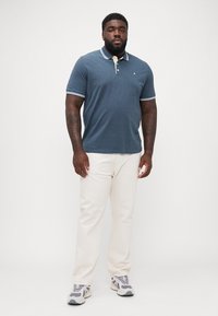 Heren blauwe polo met contrasterende witte kraag en manchetten, gecombineerd met lichtbeige broek en grijze sneakers.