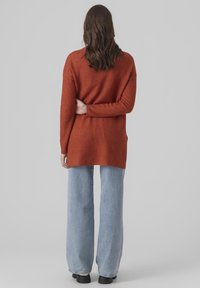 Femme aux cheveux longs bruns portant un pull orange rouille, un jean bleu clair à jambes larges et des chaussures noires, debout de dos avec le bras derrière le dos.