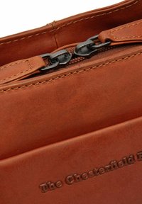 Bolso de cuero marrón con una superficie texturizada, que cuenta con un cierre de cremallera y herrajes en metal oscuro. El nombre de la marca está estampado en la parte frontal.