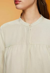 Blusa de manga larga beige claro con cuello, textura suave, detalle de pliegues en la parte frontal y un cierre de botón en el cuello.