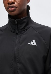 Gros plan d'une personne portant une veste noire zippée avec un logo à trois bandes blanches sur la poitrine gauche sur un fond uni.