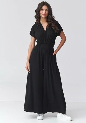 Kobieta z falowanymi włosami, ubrana w czarny kombinezon z szerokimi nogawkami, krótkimi rękawami i dekoltem w serek, w białych trampkach, stojąca z jedną ręką w kieszeni.