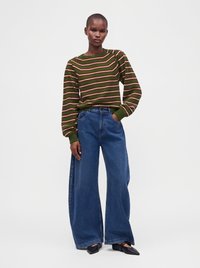 Personne portant un pull rayé vert et rouge, un jean bleu à jambes larges et des chaussures noires pointues, debout devant un fond clair uni.