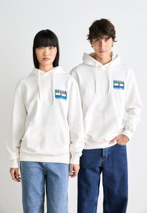 Zwei junge Erwachsene, die weiße Kapuzenpullover mit bunten Logos auf der Brust tragen, kombiniert mit blauen Jeans, stehen vor einem schlichten weißen Hintergrund.
