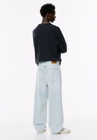 Ung mand med kort, krøllet hår iført en sort sweatshirt, lyseblå bredbenede jeans og hvide sneakers, stående med armene krydset.